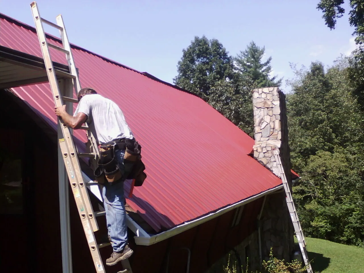 Roofer on a ladder beside a red metal roof and stone chimney — Soffit & Fascia Repair by Roofing Brothers in Clarksville